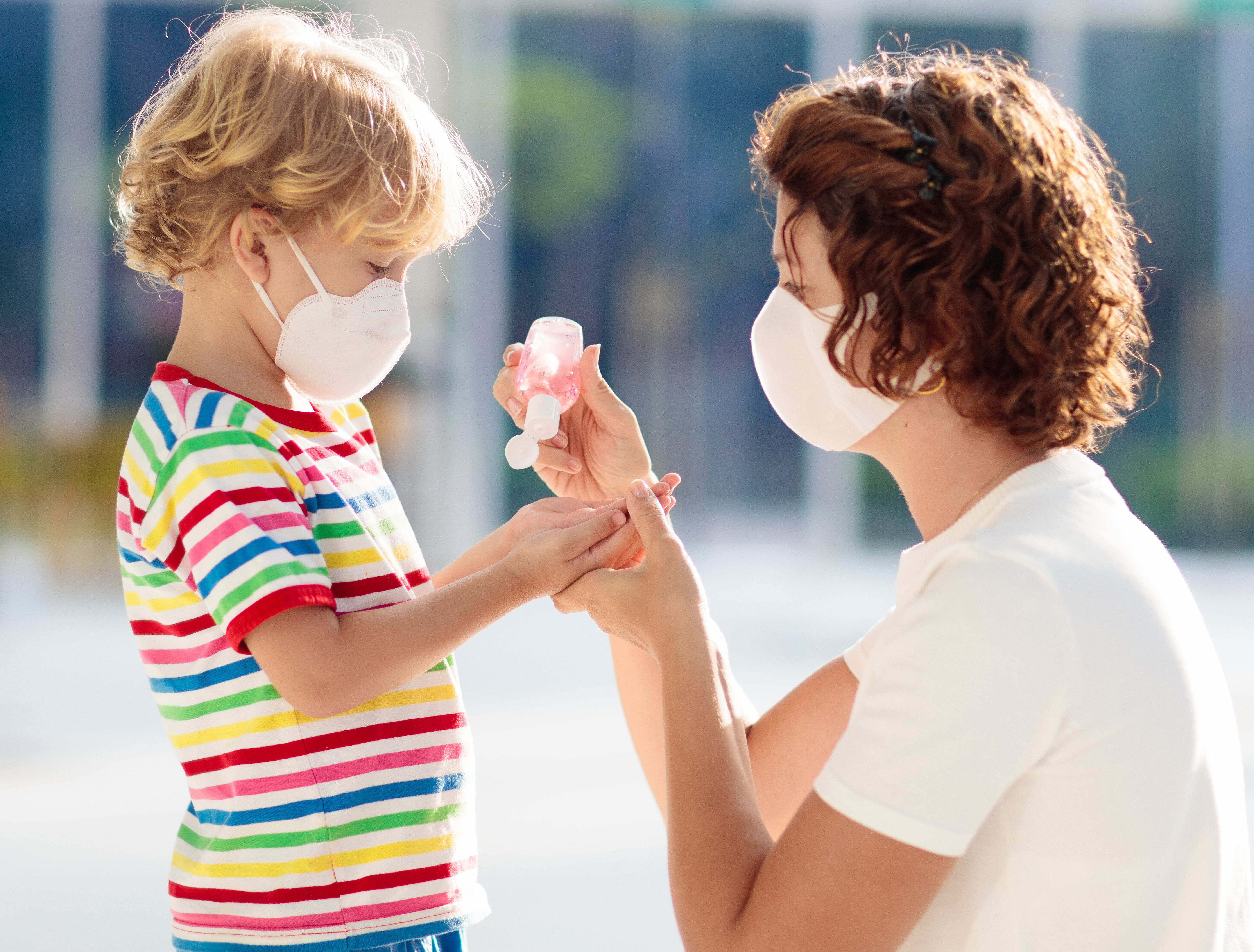 eine Mutter mit Mundschutz desinfiziert ihrem kleinen, Mundschutz tragenden Kind, die Haende